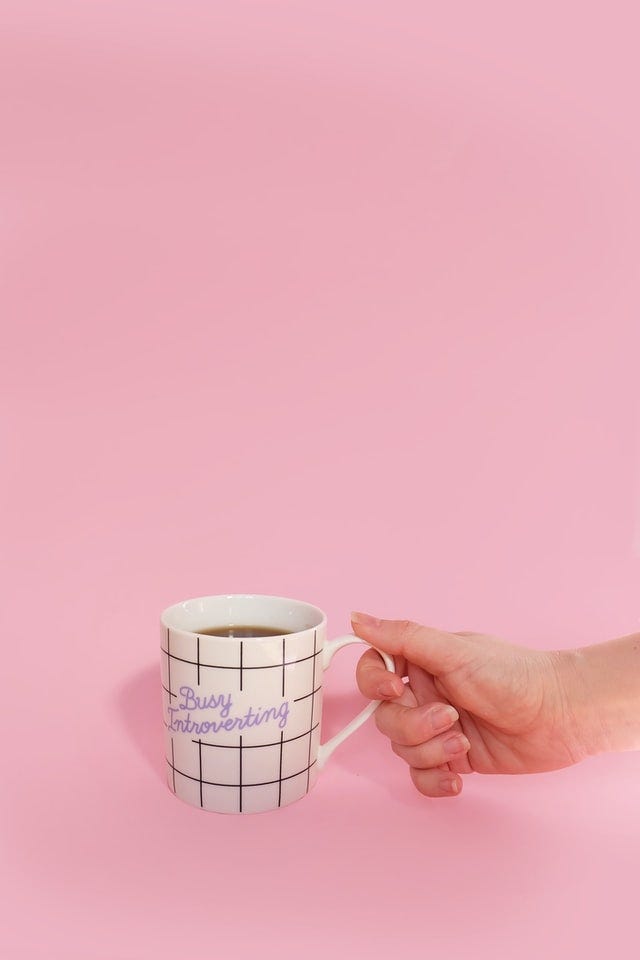 Uma mão segurando uma xícara branca com linhas pretas contendo o texto "Busy Introverting" em roxo sobre um fundo rosa pastel.