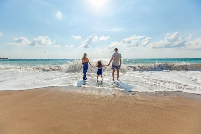 Two adults and one child standing at the water line at the beach in front of blue sky,