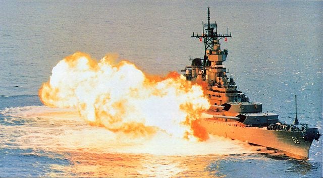 The battleship USS Missouri firing a broadside of its 16-inch guns. The ship is diagnonally positioned on the right side of the picture and the balls of flame from the guns firing dominate the center and left of the picture