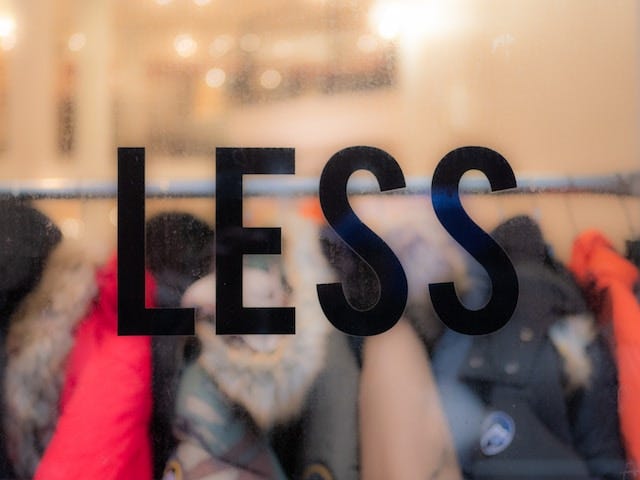The word LESS is superimposed over a blurred background of clothes hanging on a rack at a store.