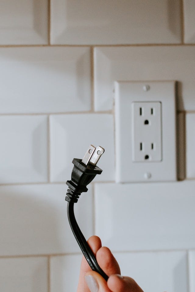 A hand holds a black two pronged plug (USA Plug) in front of a tiled wall with two socket electrical outlet.