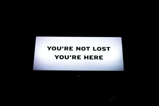 A white sign against a solid dark background. It reads: “YOU’RE NOT LOST. YOU’RE HERE.”