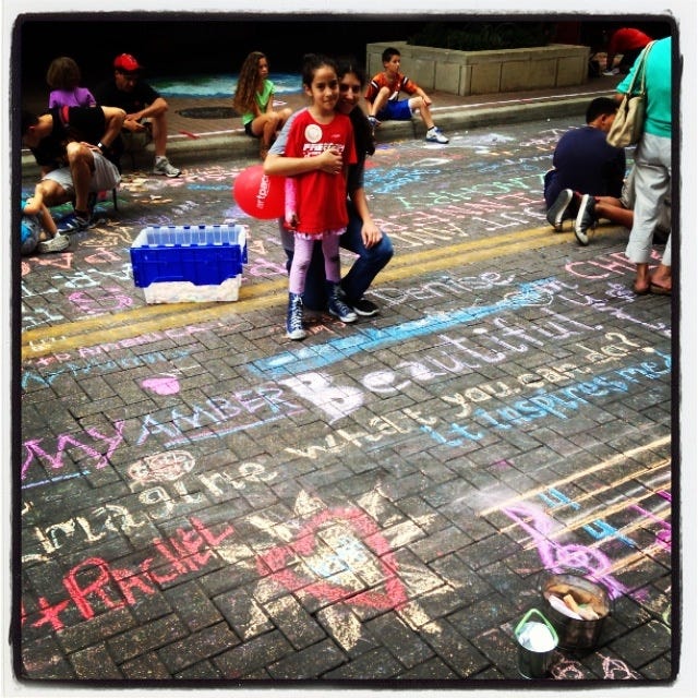 A young girl poses with a smile on a street filled with colorful chalk art and positive messages. She holds a red balloon and is surrounded by other participants engaged in creating chalk drawings. The vibrant artwork on the pavement includes uplifting words and drawings, creating a lively and creative atmosphere