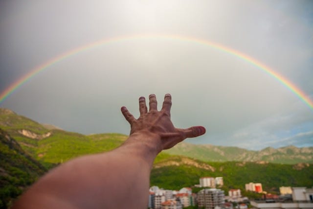 A left hand reaching out towards a rainbow, in a grey sky, over green hills and out-of-focus tall buildings in the foreground.