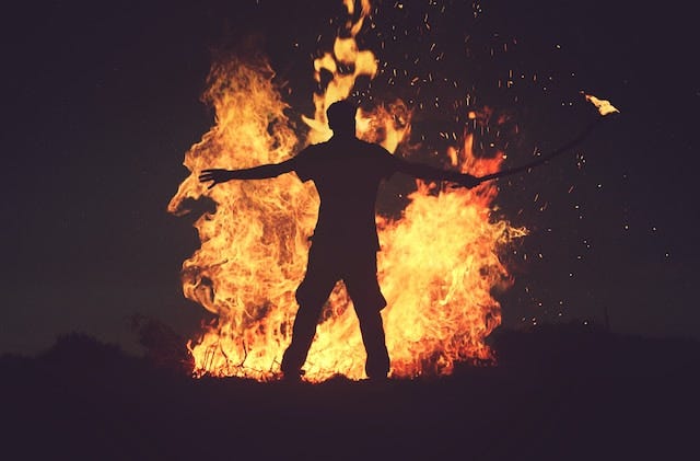 Photo of silhouetted figure in front of flames, holding a stick with fire on the end.