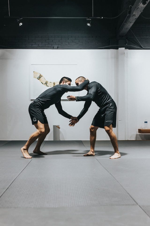 Two men standing barefoot in a grappling position. Both are wearing tight, black, long sleeve shirts, and long, black board shorts.