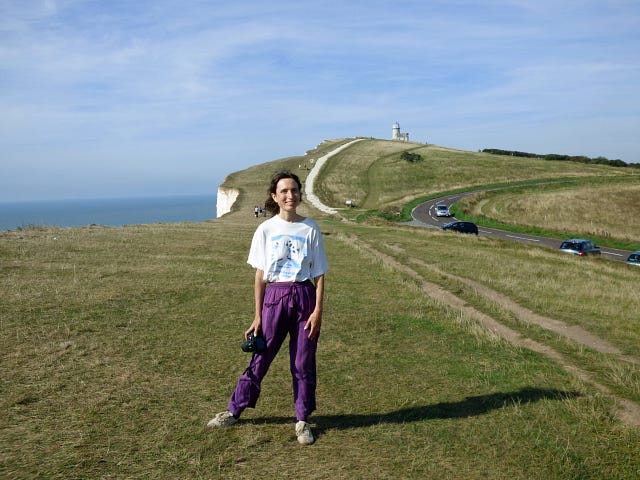 A woman, Susie Kearley, in purple trousers and a white T shirt, on grass next to a coast path, the sea in the distance, a lighthouse faintly in the distance, in Eastbourne
