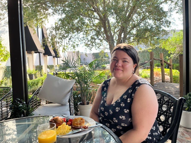 Brittany smiles at the camera at a table with breakfast in front of her and greenery in the background.