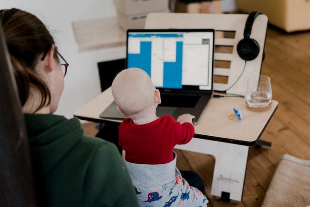 A mother working from home with her baby on her lap. The baby is curiously looking at the notes on the mother’s laptop screen. This image is used as a metaphor for mothers who are balancing work and childcare.