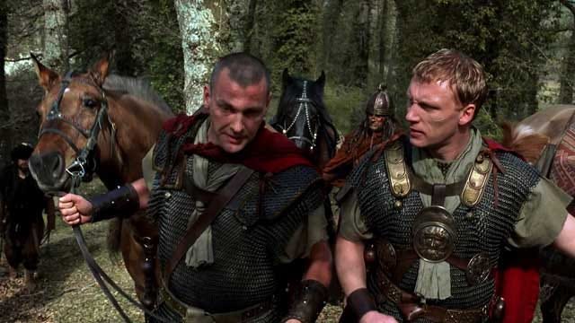 Two men in roman armour with red capes led their horses through a forest.