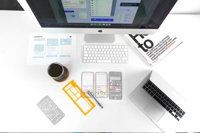 Aerial view of a desk with a monitor, keyboard laptop, stencils of phone apps, coffee cup, paper of wireframes, and a book