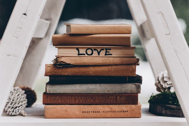 A stack of old and vintage books sits under a small, white-painted ladder. The books mostly in shades of brown. One has the pages turned forward instead of the spine, and someone has written “LOVE” on it in black marker.