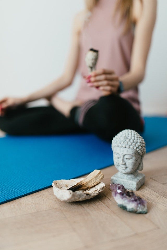 woman in pink top and black leggings sits on blue yoga mat holding burning sage, in the front there is a Buddha statue and bowl of burning Palo Santo