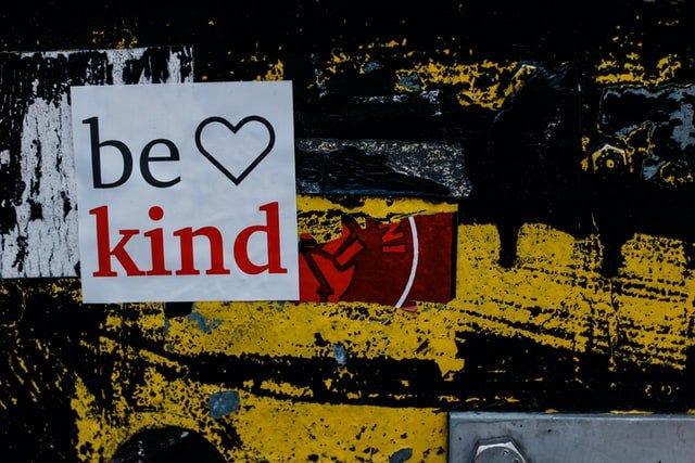 “Be Kind” sign on wall with black and yellow graffiti