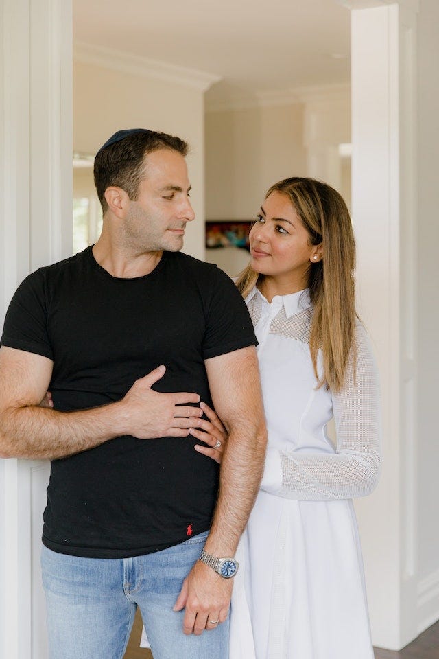 Man and woman look at each other, he is standing slightly to the front, wearing a black top, with his hand holding his partner’s hand. She stands slightly behind wearing a white dress. They look at each other with affection.