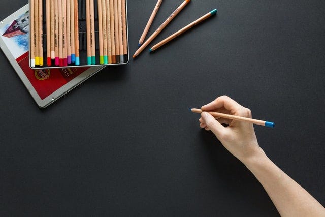 Open box of colouring pencils, some outside. Hand holding one as if writing. Black background.