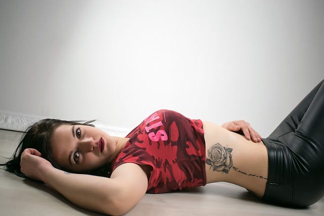 Young woman, dark hair, red cropped t-shirt, rose tattoo and black latex trousers, lies on her back & looks at camera