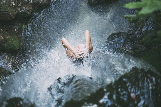 A woman showering/bathing under a waterfall surrounded by large walls of stones.