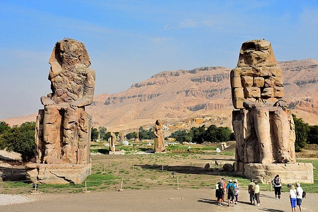 Two giant statues of an Egyptian Pharaoh outside, with people standing in front of them taking pictures.