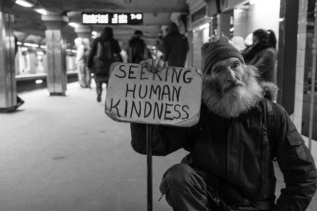 Black and white image of an older man with white beard in subway station holding sign saying “Seeking human kindness.”