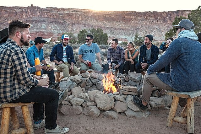 A group of happy young people gathered around a campfire. In the background some leafy trees, then further back a rocky line of cliffs with the sun low behind.