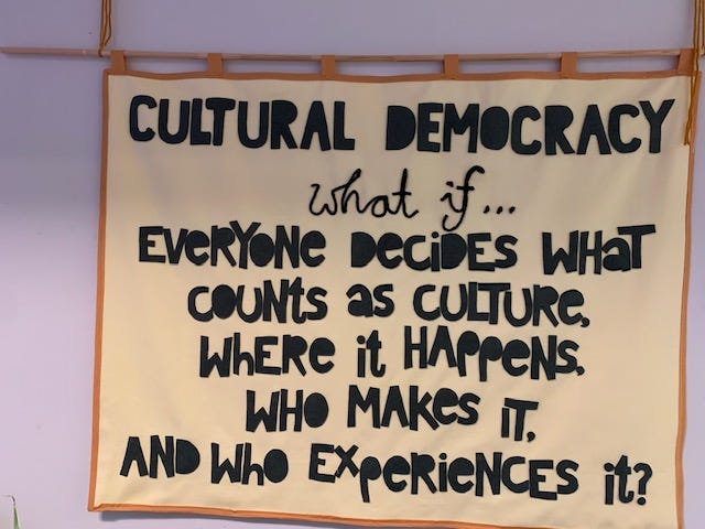 Showing a stone coloured canvas banner with the writing ‘cultural democracy what if … everyone decides what counts as culture, where it happens, who makes it and who experiences it?’ stitched on in black fabric. Banner created by Time Rebels Ruth and Odette of We Are Makers