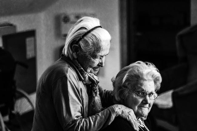 black and white portrait of two elderly women, one of whom is seated and looking at the camera, while the other is standing behind her, and gazing to the side of the frame.