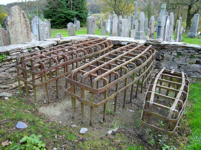 A colour photo of mortsafes in a graveyard. They look like large, rusty iron cages above graves. They look like hollow iron coffins.