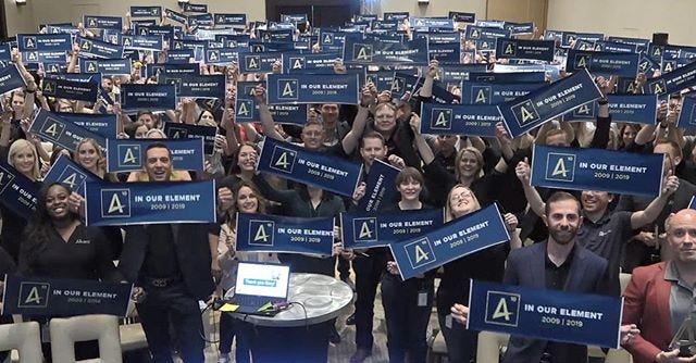 An image of a room full of Alkamists holding up banners that say “In our Element” with a date and “chemistry symbol” of an A