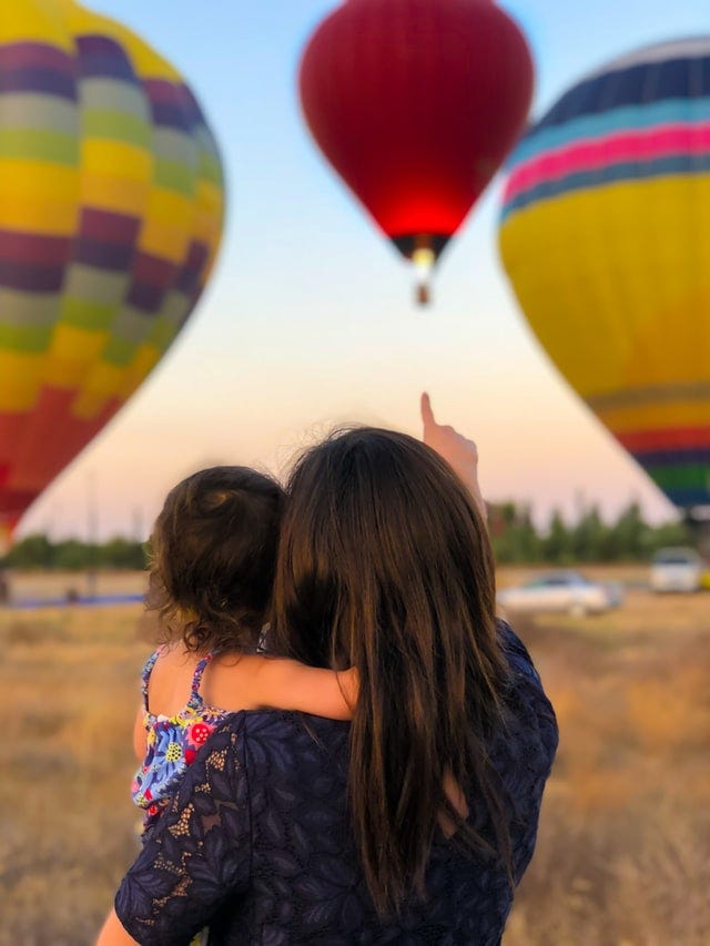 A mother carrying her toddler and pointing to big hot air balloons, rising up in the sky. Probably she is explaining to the child, why one of the balloons is going upward. This photo is used as a metaphor for the interesting conversations between a mother and her child.