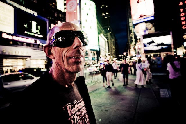 Man with sunglasses in Times Square at night. There are people and cars in the street and neon lights on the buildings.