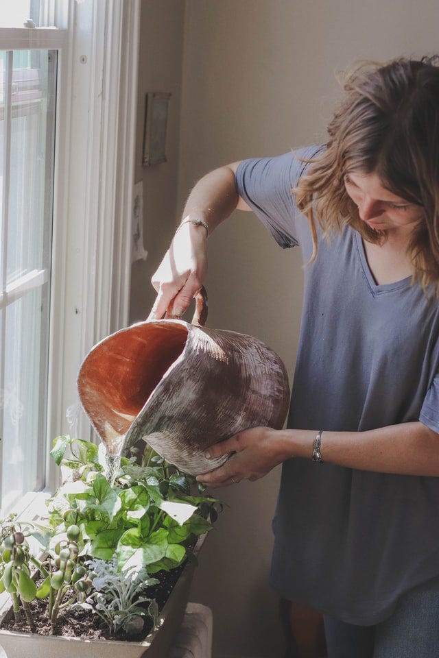 Caucasian woman in a loose blue-gray shirt waters a plants in an indoor window box with a large copper pitcher.