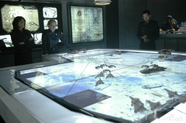 A scene from Battlestar Galactica with the characters overlooking a tactical planning table depicting the events of a battle.