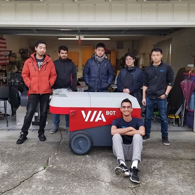 Group of 5 people standing in a garage behind a rolling robot; Akhil is sitting in front of the robot.