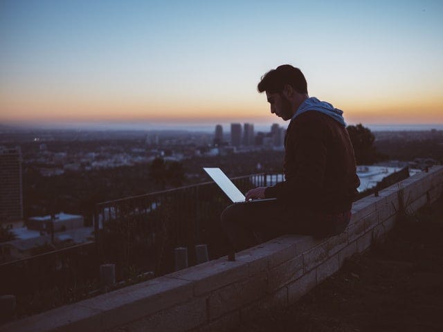 man on a ledge with his laptop at the rooftop overlooking the city sunrise