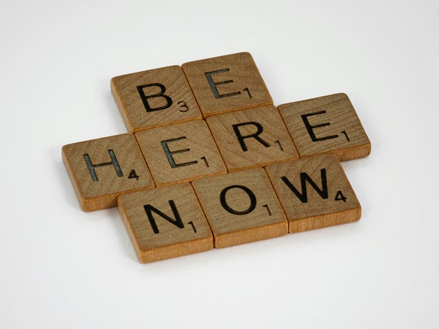 Scrabble tiles on a white background spelling out Be Here Now.