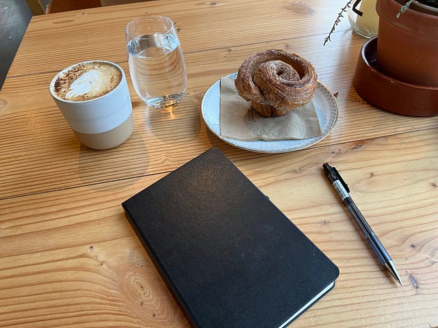 A latte, glass of water, cinnamon bun, journal and pen lay on a wood table in a coffee shop