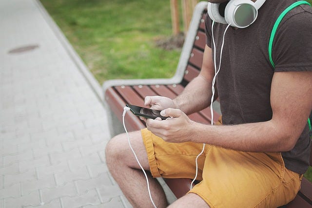 Man sitting in a bench holding a phone with plug in headphones on his neck