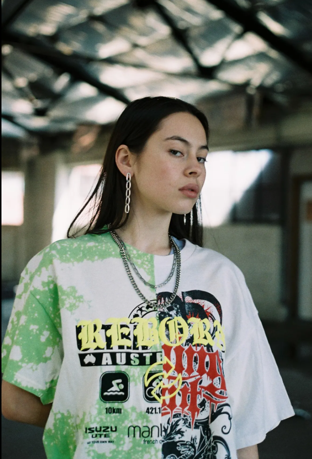 A woman stands in an empty warehouse. She is wearing a jumper with ‘Reborn’ on the front and metal jewellery.