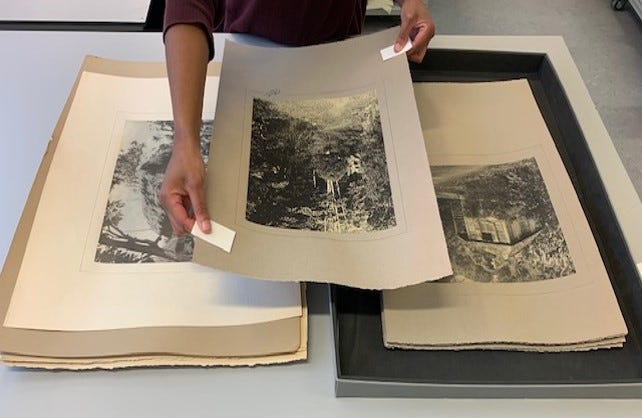 A conservator carefully lifting a print from a stack of prints at alternate corners using small strips of archival paper which have been folded around the artwork.