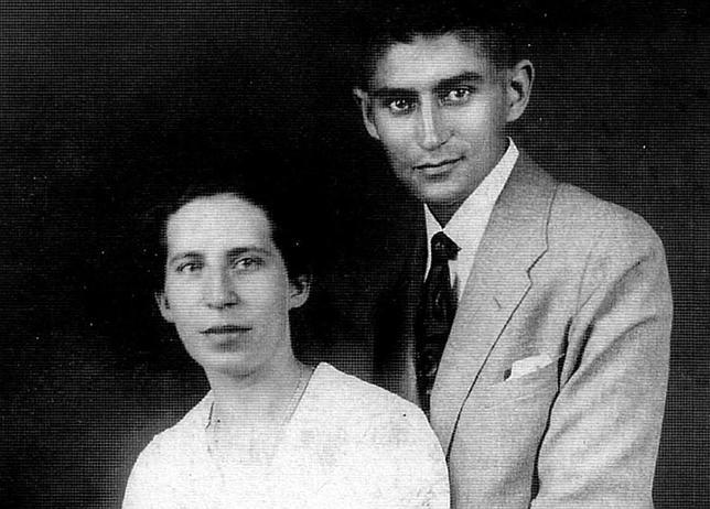 A foto mostra um homem e uma mulher posando para uma foto. O homem está vestindo um terno branco formal estilo retrô com óculos, posando ao lado de uma mulher. O cenário parece ser interno contra uma parede. O homem é Kafka e a mulher é Felice Bauer.