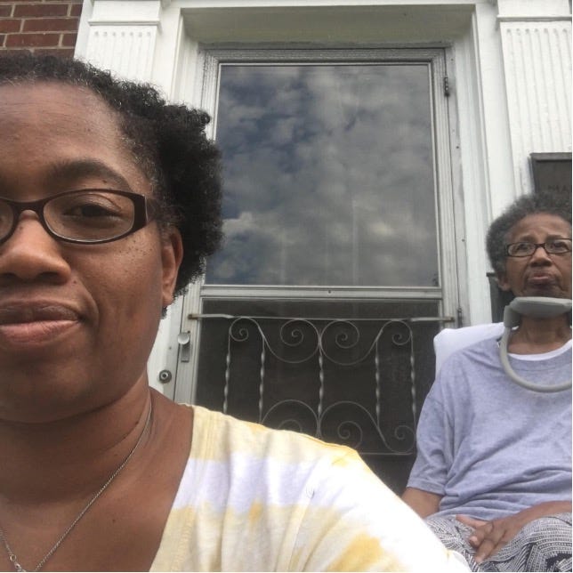 Candace with her mom on the front porch. Candace is in the foreground smiling at the camera. Her mom is sits behind her and gazes at the street.