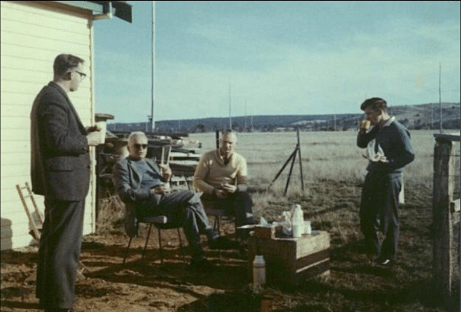 Graeme Ellis and Grote Reber outside Reber’s hut by an radio array outside Hobart in 1965, along with colleagues Bart Bok  and Peter McCullough. Bok and Reber are sitting in the center and Ellis and McCullough are standing; all are drinking from mugs around a makeshift table.