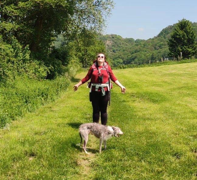 a photo of Bella standing in a field, her arms stretched out. Its very sunny, Bella is wearing sunglasses and a backpack. In front of her in a grey and white dog.