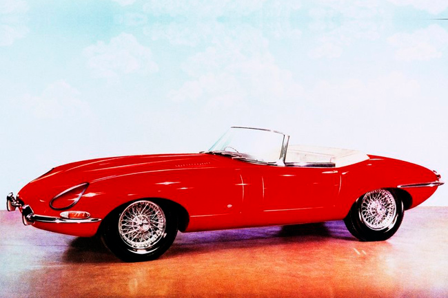 Na imagem temos o Jaguar 1961 tipo-E vermelho com um fundo em azul claro com nuvens atrás dele.