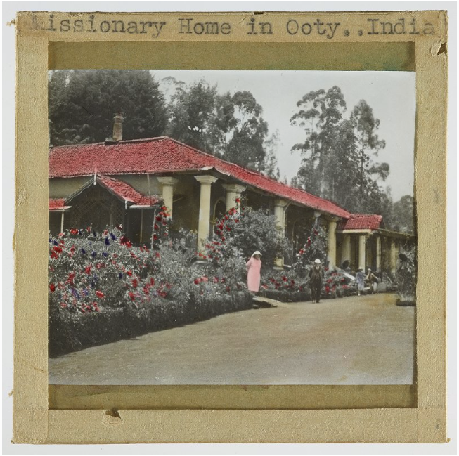 Photograph with red coloured hues of a house with red rooves, white columns and flower bushes, featuring a central figure in pink.