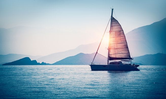 A sailing boat at sunset with a mountainous shore in the distance.