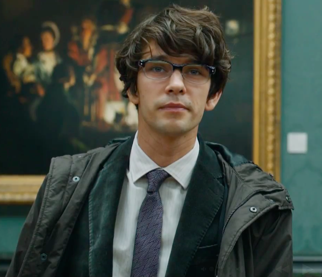 Ben Whishaw a young man with mid length hair wearing glasses and a suit and tie with coat is sitting in front of a classical painting. The painting behind depicts he is in a gallery.