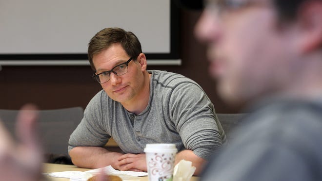 A man with light skin, glasses, and dark brown hair watches a blurred student in the foreground speak.