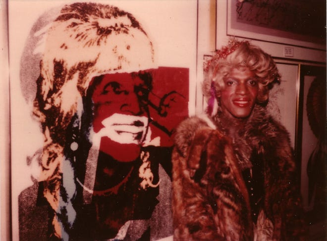 Marsha P. Johnson posando em frente ao retrato composto por Andy Warhol — veste um casaco de pele, uma peruca loira, batom.